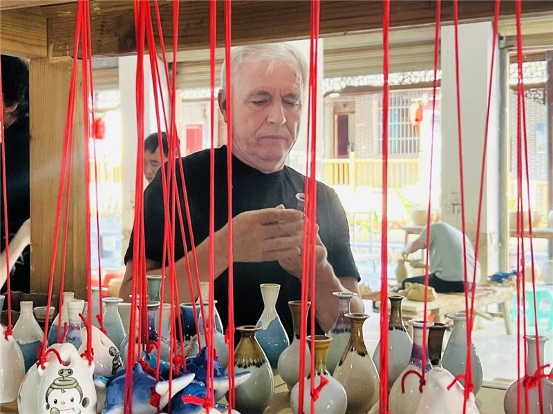 鄉(xiāng)愁如同一根根紅線，將游子與故鄉(xiāng)永遠(yuǎn)地牽連到一起?；逄账囄幕瘎?chuàng)意一條街的母氏陶藝館內(nèi)，紅線吊著陶瓶，思緒游走萬(wàn)里。摩爾多瓦“信息標(biāo)簽”通訊社社長(zhǎng)亞歷山大·塔納斯端詳著館內(nèi)展示的陶藝作品，并挑選出心儀的小擺件帶回摩爾多瓦，也帶走花茂村的“鄉(xiāng)愁”。（張亞欣攝）